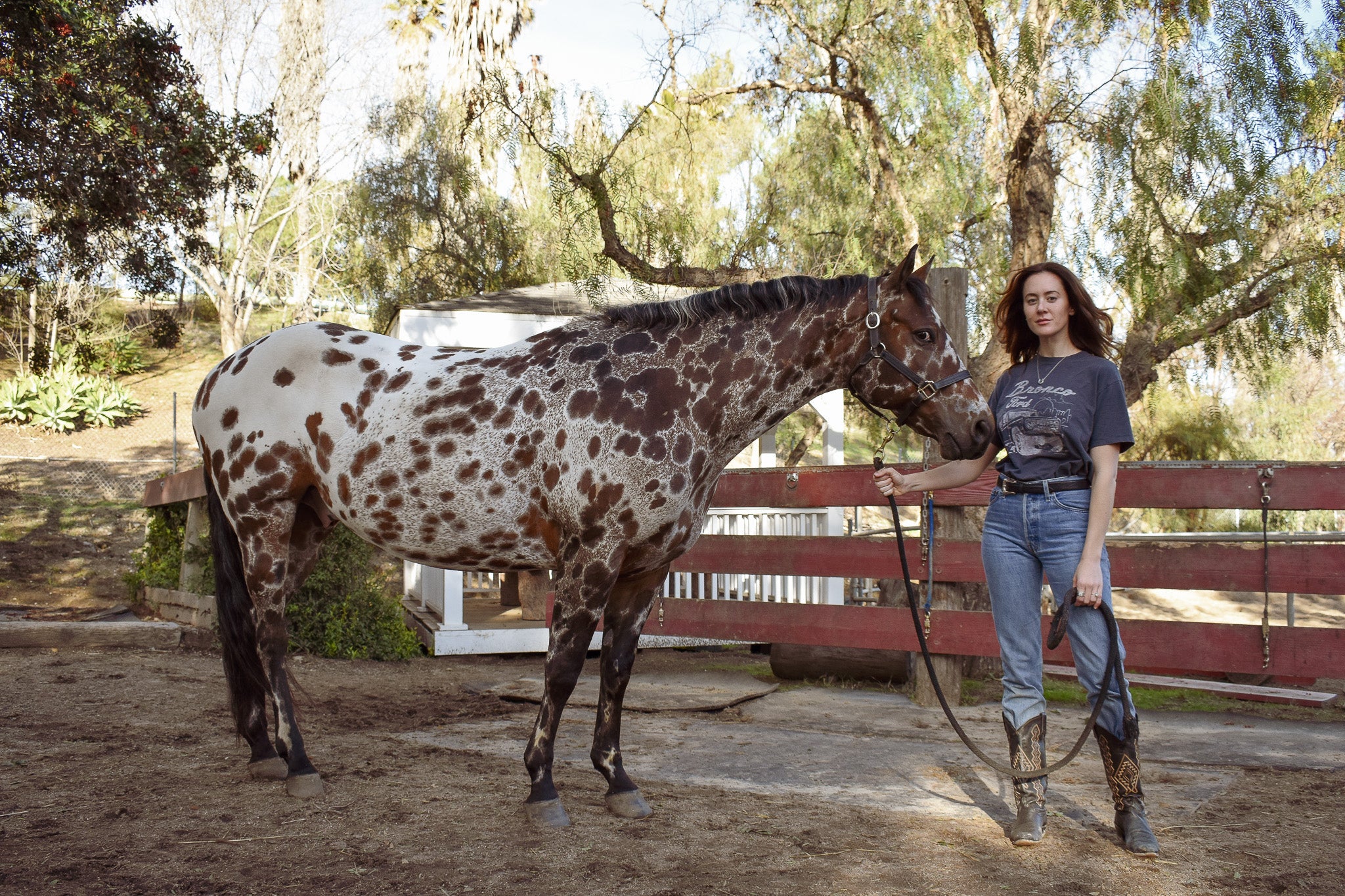 Model standing strong and confident next to an Appaloosa horse, wearing the horseshoe pendant in country western wear for Saddle & Stone